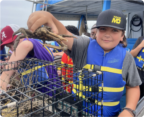 Boy in blue life jacket and cap holding a crab above a wire trap on a boat